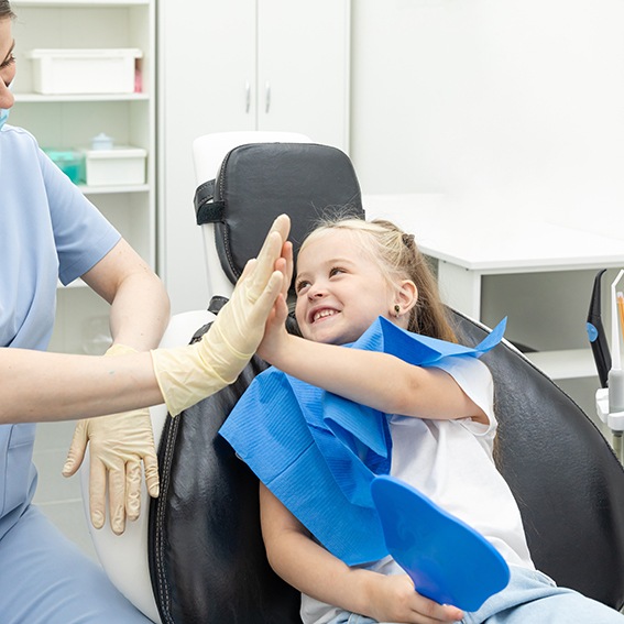 Little girl in dental chair high-fiving dentist in blue scrubs