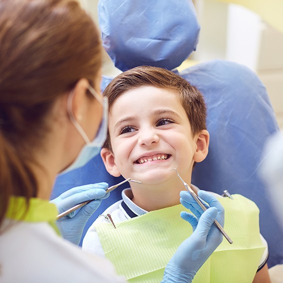 Litte boy in dental chair smiling as dentist approaches with special instruments