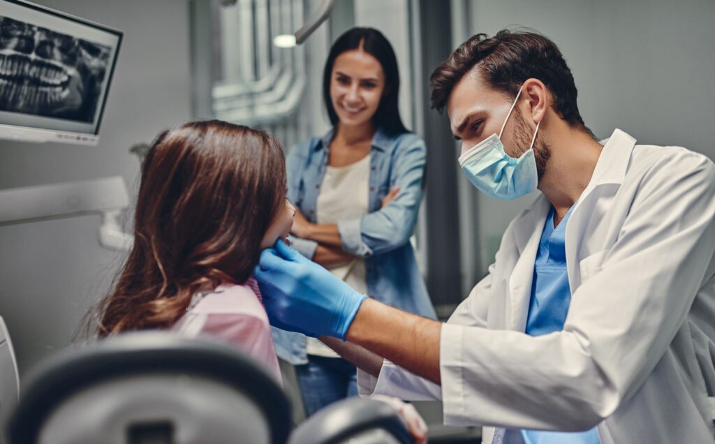 Dentist in blue gloves examining girl's teeth with mom in background