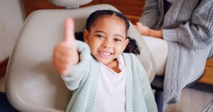 Little girl in dental chair giving thumbs up with parent in background
